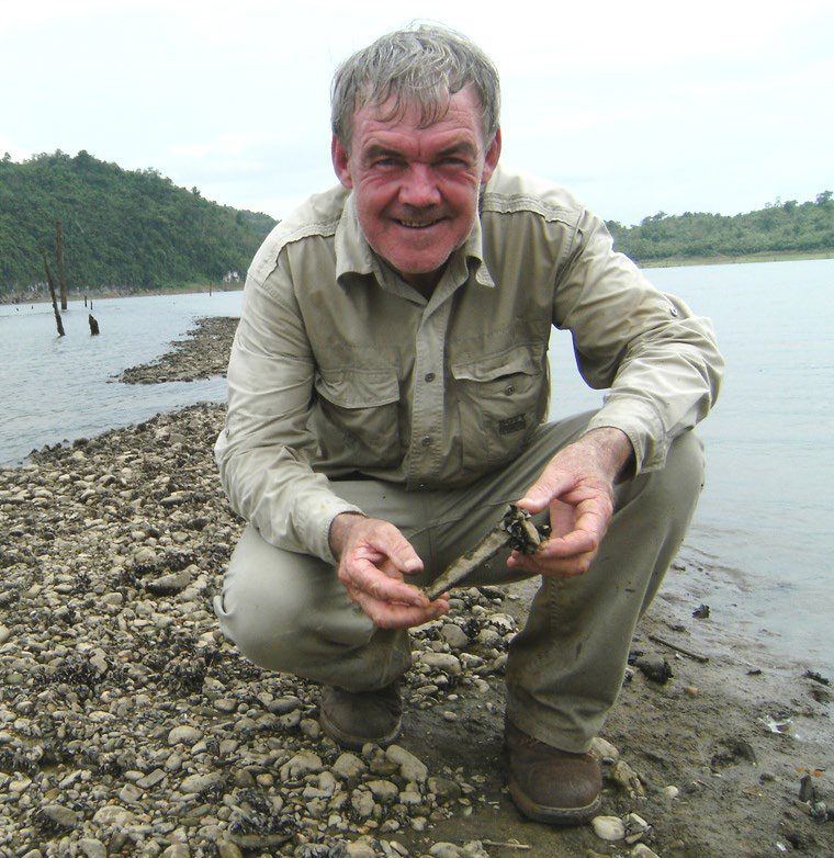 Rod Beattie crouching on a stony shoreline holding a railway spike